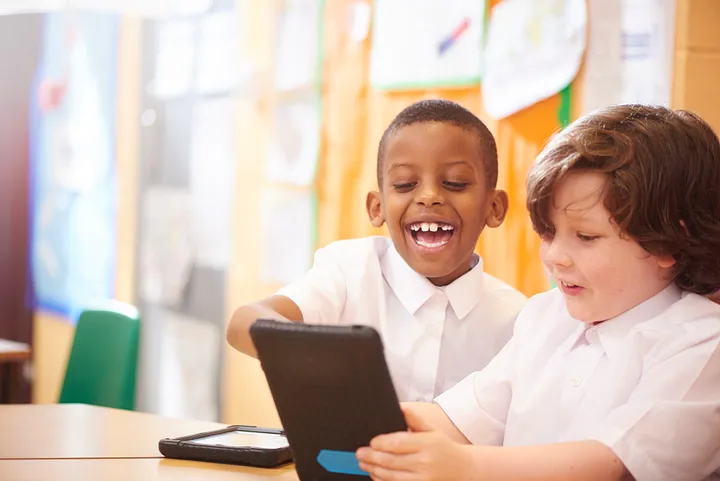 Two children in a classroom setting are seated at a table, engaging with a tablet device. 