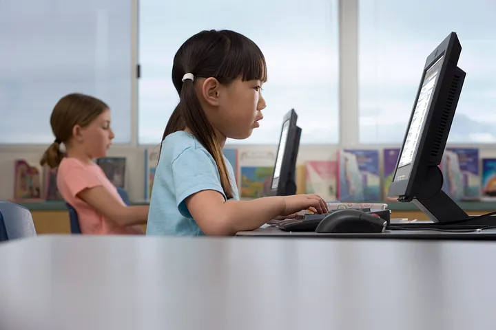 Two children sitting at desks using desktop computers in a classroom setting, with bookshelves and windows visible in the background.