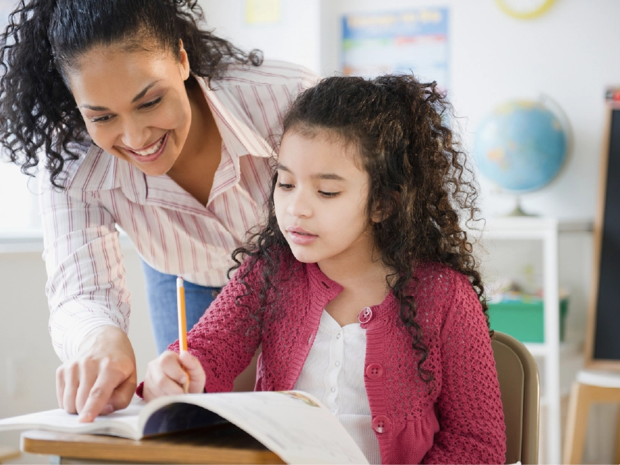 A student at a desk writes in a workbook while her teacher points at the text while smiling.